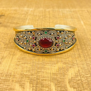 May include: A gold-toned bracelet with intricate filigree detailing and a large red gemstone in the center. The bracelet is adorned with smaller green and red gemstones.