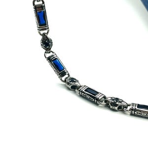 Può includere: Una collana argentata con maglie rettangolari e ovali alternate. Le maglie rettangolari presentano una gemma blu vibrante, mentre le maglie ovali hanno una pietra blu più scura. Il design presenta un motivo ornato e dettagliato.