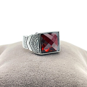 May include: A silver ring with a large, square, faceted, deep red gemstone. The ring has a geometric design on the sides. The ring is displayed on a gray surface. Jewelry.