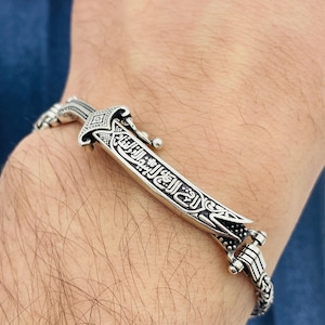 Puede incluir: Una pulsera de plata con un colgante de espada detallado. La espada tiene grabados intrincados y una hoja curva. La pulsera está hecha de piezas de plata enlazadas, creando una banda flexible. El diseño es ornamentado.