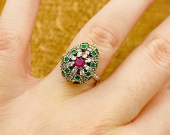Può includere: Un anello d'oro con un design a forma di lacrima che presenta una grande pietra rossa circondata da pietre bianche e verdi più piccole.