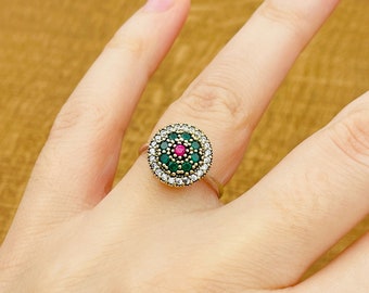 Può includere: Un anello d'oro con un designo rotondo che presenta una pietra centrale rosa circondata da pietre verdi e un bordo di pietre bianche.