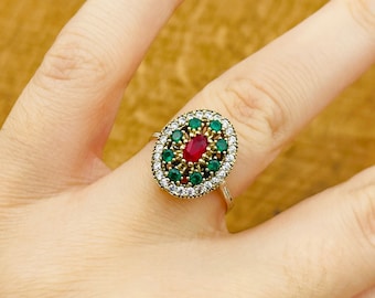 Può includere: Un anello d'oro con una pietra ovale rossa circondata da pietre verdi e bianche.