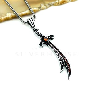 Op de afbeelding: Een zilveren hangerketting met een gebogen zwaardontwerp. Het zwaard heeft een zwarte handgreep en lemmet, versierd met rode edelstenen en een oranje middensteen. De ketting is ook zilver.