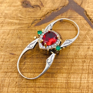 May include: A silver ring with a red pear-shaped stone surrounded by smaller clear stones. The ring has two bands that are connected by a hinge. The stone is accented with two green stones on either side.