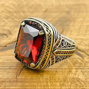 May include: A silver ring featuring a large, square-shaped red gemstone surrounded by smaller brown gemstones. The ring band has intricate gold-coloured detailing.