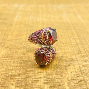 May include: A silver ring with two large red gemstones surrounded by smaller red gemstones. The ring is on a wooden surface.