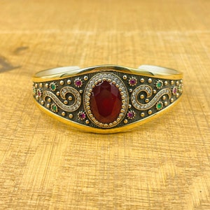 Puede incluir: Una pulsera de puño dorada con una piedra preciosa roja ovalada y facetada en el centro. La pulsera presenta un diseño detallado con pequeñas piedras preciosas rojas, verdes y transparentes, y un patrón de filigrana plateada. La pulsera se muestra sobre una superficie marrón claro.