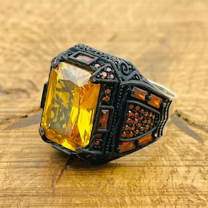 Op de afbeelding: Een zwarte ring met een grote, gele, gefacetteerde edelsteen gezet in een gedetailleerd, versierd ontwerp. De ring is versierd met kleinere, rode edelstenen.