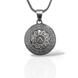 May include: A circular silver-toned pendant on a metallic chain. The pendant features a central Greek Lambda symbol surrounded by a starburst pattern and a detailed Greek key border. The metal has an oxidised finish with dark recessed areas.