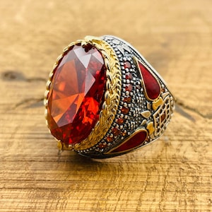 May include: A silver ring with a large, red, faceted gemstone set in a gold-toned bezel. The ring is decorated with intricate details and smaller red gemstones.