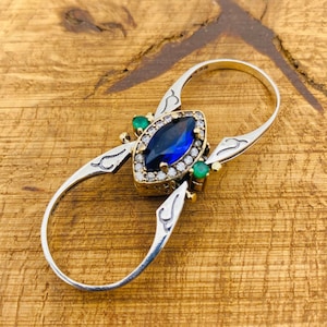 Könnte beinhalten: Ein silberner Ring mit einem blauen, markisenförmigen Edelstein, der von kleineren, klaren Edelsteinen umgeben ist. Der Ring hat auf jeder Seite des Mittelstücks zwei grüne Edelsteine. Der Ring ist so konzipiert, dass er an zwei Fingern getragen werden kann.