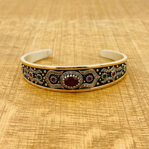 Può includere: Un braccialetto rigido in argento con un design in smalto nero e pietre preziose rosse e verdi. Il braccialetto ha un design delicato e intricato.