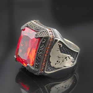 Peut inclure: Une bague en argent avec une grande pierre rouge et des détails complexes. La bague présente un motif stylisé sur la bande, avec de petites pierres serties autour de la pierre précieuse.