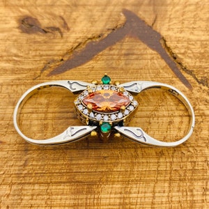 Könnte beinhalten: Ein silberner Ring mit einem großen orangefarbenen Edelstein, der von kleineren weißen Edelsteinen umgeben ist. Der Ring hat zwei Bänder, die durch ein dekoratives Design mit grünen Edelsteinen verbunden sind.