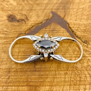 Könnte beinhalten: Ein silberner und goldener Ring mit einem schwarzen und weißen Edelstein in der Mitte. Der Ring ist im Doppel-Loop-Stil gestaltet, wobei der Edelstein in einem dekorativen Rahmen gefasst ist.