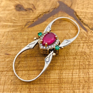 May include: A silver ring featuring a red pear-shaped gemstone surrounded by smaller clear gemstones. The ring has two bands connected by a decorative design. The ring is accented with two green gemstones.