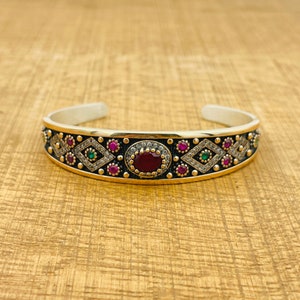 Può includere: Un braccialetto d'argento con un designo nero e oro. Il braccialetto è decorato con piccole pietre rosa, verdi e bianche. Una pietra rossa più grande è al centro del designo.