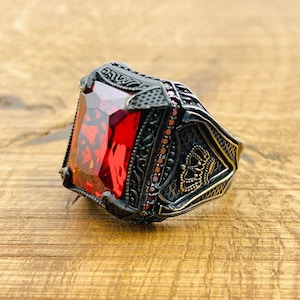 Könnte beinhalten: Ein silberner Ring mit einem großen, quadratischen, roten Edelstein. Der Ring ist mit komplizierten, detaillierten Designs und einer Krone an der Seite verziert.