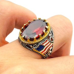 Op de afbeelding: Een zilveren ring met een grote ovale, dieprode edelsteen. De ring heeft een goudkleurige kroonzetting rond de steen en een Amerikaans vlagontwerp aan de zijkant. De ring wordt aan een vinger gedragen.