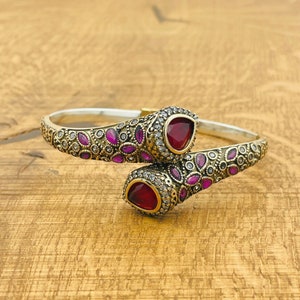 May include: A silver and gold bangle bracelet with intricate floral designs and two red gemstone accents.