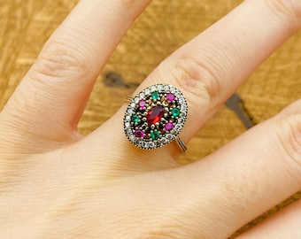 Può includere: Un anello d'argento con una pietra ovale rossa circondata da pietre più piccole di colore verde e rosa. L'anello è incastonato in un metallo dorato.
