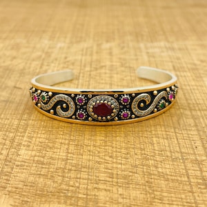 Peut inclure: Un bracelet jonc en argent avec une finition oxydée noire. Le bracelet est décoré d'un motif en spirale et présente des pierres précieuses rouges et vertes.