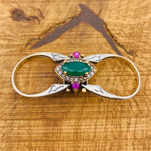 Könnte beinhalten: Ein silberner Ring mit einem grünen Edelstein, der von kleineren klaren Steinen und zwei rosa Steinen umgeben ist. Der Ring ist so konzipiert, dass er an zwei Fingern getragen werden kann.