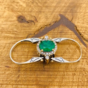 Könnte beinhalten: Ein Silber- und Goldring mit einem großen grünen Edelstein, der von kleineren weißen Edelsteinen umgeben ist. Der Ring ist so konzipiert, dass er an zwei Fingern getragen werden kann.