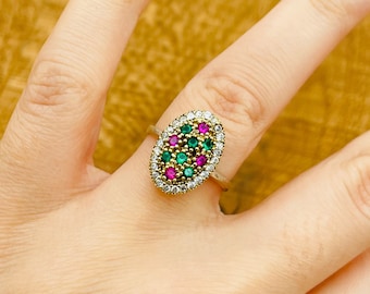 Può includere: Un anello d'oro con una montatura ovale che presenta un gruppo di pietre preziose verdi e rosa circondate da pietre trasparenti più piccole.
