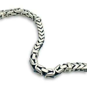 May include: Close-up of a silver necklace with a detailed, interlocking chain design. The necklace has a textured, geometric pattern, and is set against a white background. The metal has a polished, reflective finish, highlighting the intricate craftsmanship.