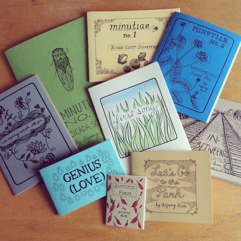 May include: A collection of small, colorful books with various titles and illustrations. The books are arranged in a circle on a wooden surface. The titles include "Minutiae No. 1", "Minutiae No. 2", "First Song", "Genius (Love)", "Autumn Poem", "Let's Go to the Park", "Cicada Song", and "In Between".