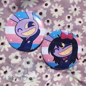 May include: Two circular pinback buttons featuring cartoon characters with purple skin and wide smiles. The left button shows a character with rabbit ears, while the right button shows a character with black hair and a red bow, both set against a background of horizontal light blue, pink, and white stripes.