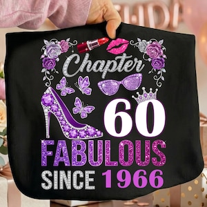 Peut inclure: Un t-shirt noir avec une impression graphique portant le texte Chapter 60 Fabulous Since 1966. Le motif comprend un escarillon à paillettes violettes, un rouge à lèvres rouge, des lunettes à paillettes violettes, des papillons, des roses et une couronne.