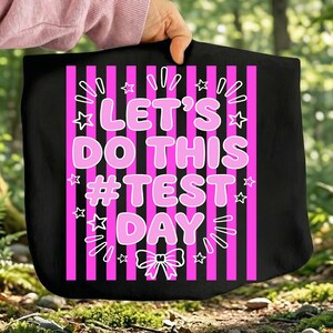 Peut inclure: Sac fourre-tout noir avec un fond à rayures roses et blanches. Le texte "LET'S DO THIS #TEST DAY" est en lettres roses à bulles. Des étoiles blanches et des lignes décoratives entourent le texte. Le sac est tenu par une personne.