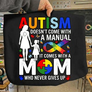 Puede incluir: Bolso tote negro con el texto "EL AUTISMO NO VIENE CON UN MANUAL, VIENE CON UNA MAMÁ QUE NUNCA SE RINDE." El diseño incluye una cinta de arcoíris, piezas de rompecabezas, mariposas y siluetas de una madre y un niño.