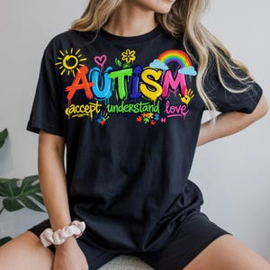 May include: Black t-shirt with the word "AUTISM" in colorful graffiti-style lettering, accompanied by the phrases "accept, understand, love" and various related graphics, including a rainbow and puzzle pieces.
