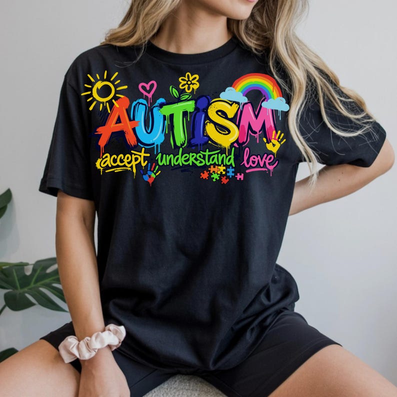 May include: Black t-shirt with the word "AUTISM" in colorful graffiti-style lettering, accompanied by the phrases "accept, understand, love" and various related graphics, including a rainbow and puzzle pieces.