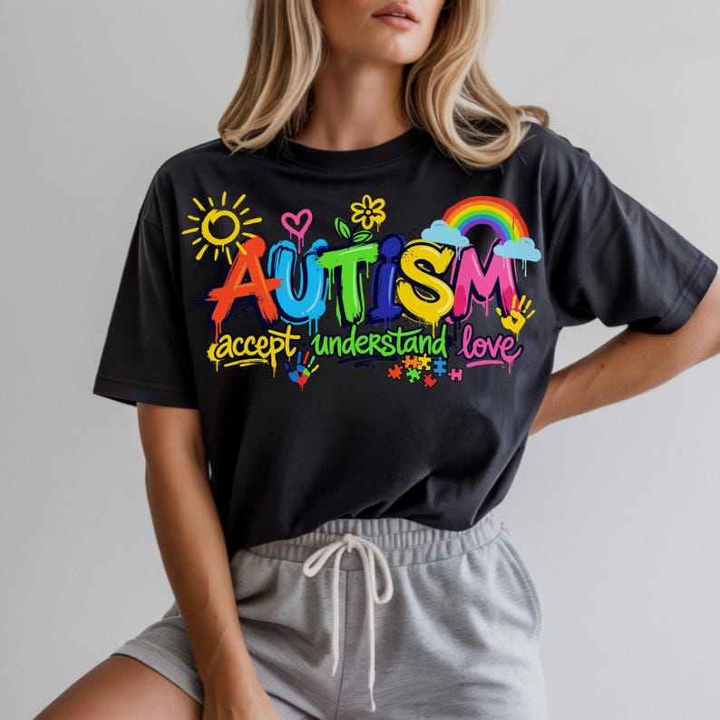 May include: Black t-shirt with a colorful graphic design. The design includes the word "AUTISM" in vibrant letters, a rainbow, sun, hearts, and the words "accept understand love". The shirt is paired with gray shorts.