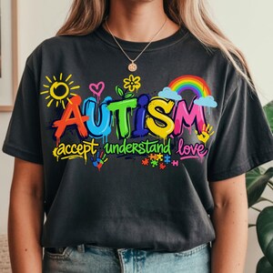 May include: Dark gray t-shirt with the word "AUTISM" in colorful graffiti-style lettering. The shirt also features the words "accept understand love" and various related graphics, including a rainbow, puzzle pieces, and handprints.