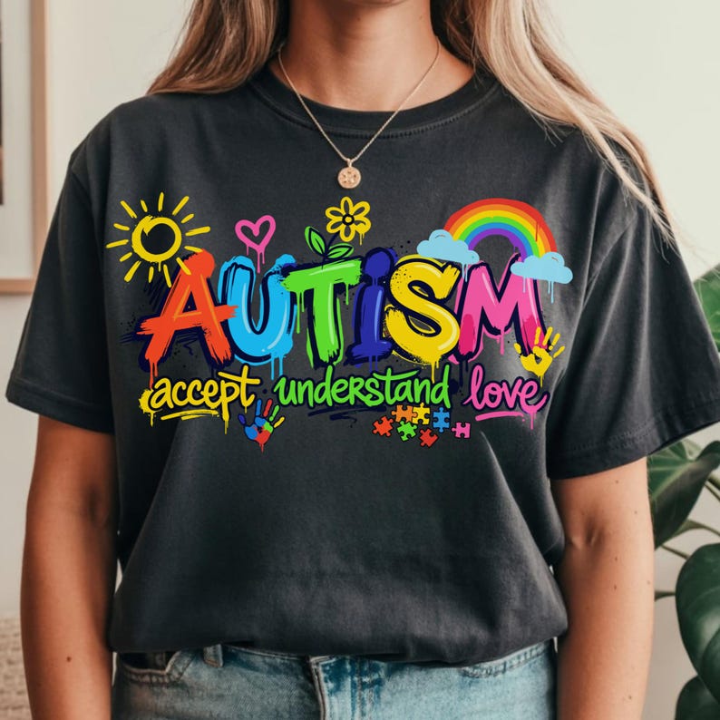 May include: Dark gray t-shirt with the word "AUTISM" in colorful graffiti-style lettering. The shirt also features the words "accept understand love" and various related graphics, including a rainbow, puzzle pieces, and handprints.