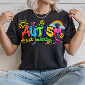 May include: Black t-shirt with a colorful graphic that reads "AUTISM accept understand love." The design includes a rainbow, sun, hearts, flowers, handprints, and puzzle pieces. The shirt is a crew neck.