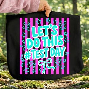 Peut inclure: Sac fourre-tout noir avec un design vibrant. Il présente des rayures roses verticales et le texte "LET'S DO THIS #TEST DAY" en turquoise. Des étoiles et un nœud turquoise complètent le design.