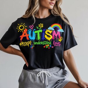 May include: Black t-shirt with a colorful graphic design. The design includes the word "AUTISM" in vibrant graffiti-style lettering, accompanied by the phrases "accept understand love" and various related symbols like a rainbow, puzzle pieces, and handprints.