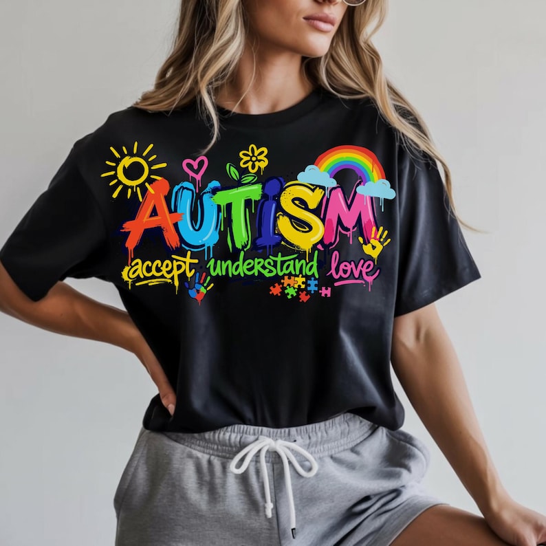 May include: Black t-shirt with a colorful graphic design. The design includes the word "AUTISM" in vibrant graffiti-style lettering, accompanied by the phrases "accept understand love" and various related symbols like a rainbow, puzzle pieces, and handprints.