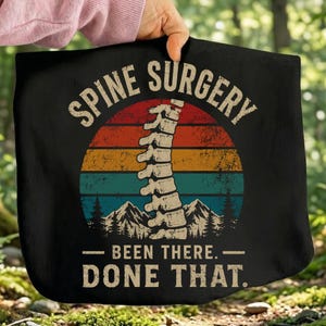 May include: A black fabric item featuring a graphic of a human spine centered over a sunset-colored circle with mountain silhouettes. The text reads "SPINE SURGERY" at the top and "BEEN THERE. DONE THAT." at the bottom in a bold, distressed font.