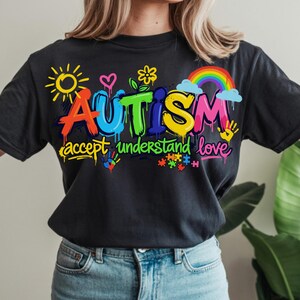 May include: Black t-shirt with a colorful graphic design. The word "AUTISM" is displayed in vibrant, graffiti-style lettering, accompanied by the phrases "accept understand love" and related imagery, including a rainbow, puzzle pieces, and handprints.