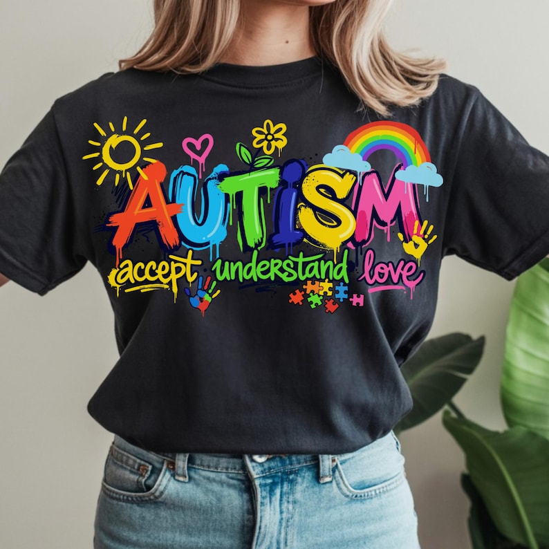 May include: Black t-shirt with a colorful graphic design. The word "AUTISM" is displayed in vibrant, graffiti-style lettering, accompanied by the phrases "accept understand love" and related imagery, including a rainbow, puzzle pieces, and handprints.