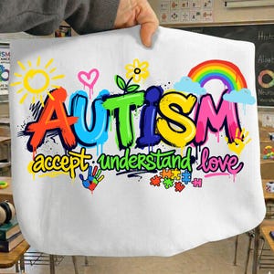 Può includere: Tessuto bianco con la parola "AUTISMO" in lettering colorato in stile graffiti. Il design include un arcobaleno, un sole, un cuore e pezzi di puzzle, con le parole "accettare, capire, amare" sotto.
