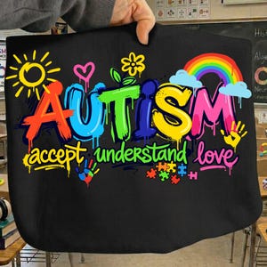 May include: Black tote bag with the word "AUTISM" in colorful graffiti-style lettering. The design includes a rainbow, sun, hearts, flowers, handprints, and the words "accept, understand, love."